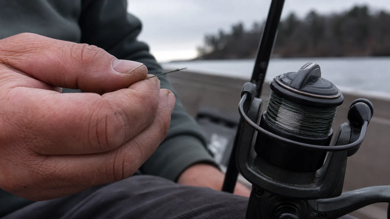 A hand holding out a broken end of fishing line next to an empty spinning reel spool on a boat deck